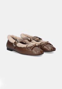 Zapatos planos de cuero marrón con forro de pelo sintético beige, correa ajustable con acentos metálicos y detalles de lazo en la parte delantera.