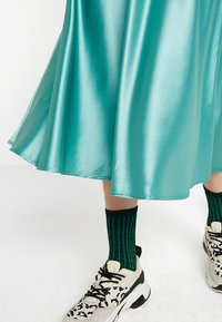 Tyrkysově modrá saténová sukně s rozšířeným lemem, nošená s černo-bílými teniskami s potiskem a zelenými žebrovanými ponožkami. Viditelná měkká, lesklá textura.