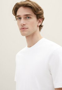 T-shirt bianca a maniche corte realizzata in tessuto liscio e leggero. Scollo rotondo con dettagli di cucitura minimale. Il modello ha i capelli spettinati e uno sfondo neutro.