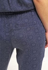 Primer plano de pantalones azul marino con sutiles patrones florales circulares y un bolsillo de welt trasero, usados por una persona con el brazo descansando al lado.