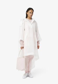 Overmaatse doorzichtige roze poncho met capuchon, gecombineerd met lichtroze broeken en witte sneakers. Gestreepte tote bag in roze en witte kleuren.