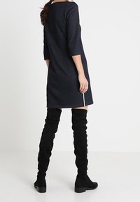 Robe bleu marine, longueur genou, avec des manches trois-quarts, dotée d'un décolleté rond et d'une fente sur le côté. Portée avec des bottes noires au-dessus du genou.