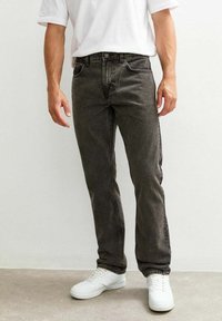Una persona sta in piedi indossando una maglietta bianca e jeans grigi scuri, abbinati a sneaker bianche. Lo sfondo è chiaro e minimalista.