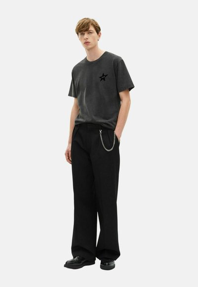 Joven con una camiseta negra de estrellas desgastada, pantalones anchos negros con una cadena plateada y zapatos de cuero negros, de pie con las manos en los bolsillos.