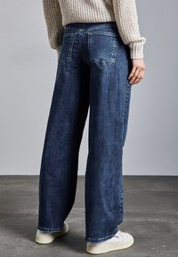 Pantalones de mezclilla oscuros de pierna ancha con una cintura de altura media, que cuentan con dos bolsillos traseros y un acabado limpio. Se llevan con zapatillas blancas.