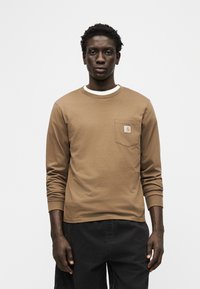 Άνδρας που φοράει καφέ πουκάμισο με μακριά μανίκια και τσέπη πάνω από ένα λευκό μπλουζάκι και μαύρο παντελόνι, στέκεται μπροστά σε ένα απλό λευκό φόντο.