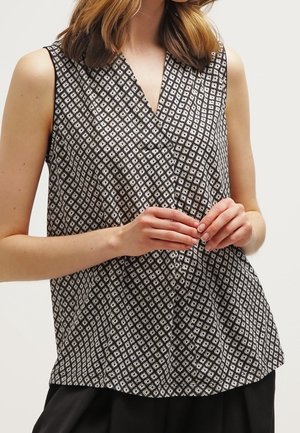 Vrouw draagt een mouwloze zwart-wit gebloemde blouse met een V-hals, staat met haar handen zachtjes gevouwen voor zich.