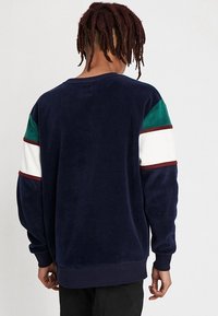 Sudadera de forro polar azul marino con puños acanalados. Presenta mangas en bloques de color verde, blanco y granate. Textura suave y ajuste relajado.