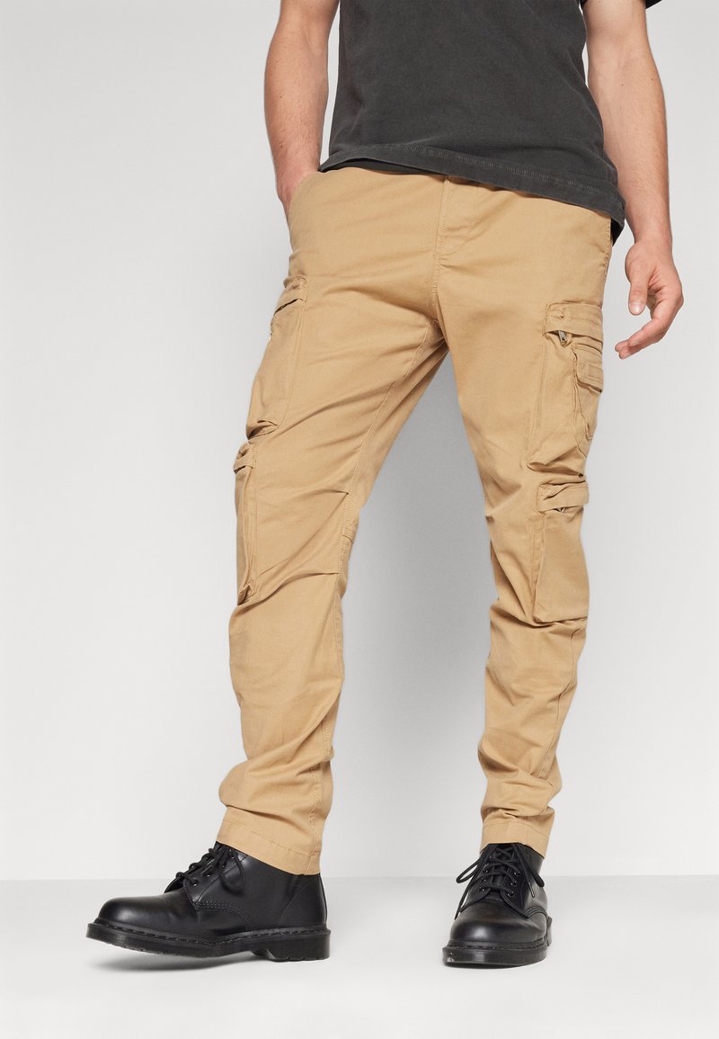 Beige Cargo-Hosen mit mehreren Seitentaschen, aus strapazierfähigem Stoff. Eng an der Taille, zulaufend zu den Knöcheln. Schwarze Schnürstiefel.