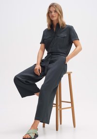 Dunkelgrauer, kurzärmliger Jumpsuit mit zwei Brusttaschen und einem passenden Gürtel. Weit geschnitten. Modell trägt hellgrüne Sandalen und sitzt auf einem Hocker.