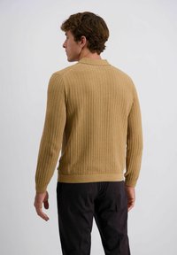 Beige gerippter Pullover mit Polo-Kragen, langen Ärmeln und taillierter Säume. Getragen über dunklen Hosen, die den strukturierten Stoff und die maßgeschneiderte Passform zur Geltung bringen.
