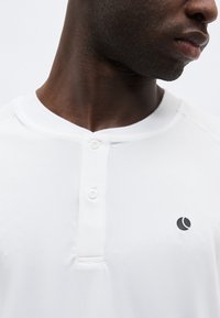 Κοντινό πλάνο του λαιμού και του ώμου ενός άνδρα που φοράει λευκό μπλουζάκι Henley με δύο κουμπιά και ένα μικρό μαύρο λογότυπο στο στήθος.