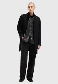Svart ullfrakk over en svart lærskjorte, kombinert med vide svarte jeans. Outfiten har et minimalistisk design og glatte teksturer.