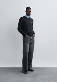 Črn pleten pulover preko svetlo modre srajce s kratkimi rokavi, oblečen z sivimi kavbojkami in črnimi čevlji. Enostaven dizajn, minimalni poudarki.