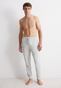 Grijze katoenen loungebroek met een elastische tailleband en een trekkoord. Aansluitend ontwerp met taps toelopende benen. Model staat blootsvoets en zonder shirt.