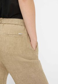 Beżowe lniane spodnie o luźnym kroju, wyposażone w boczne kieszenie oraz tylną kieszeń z srebrnym detalem. Materiał wydaje się miękki i o wyraźnej fakturze.