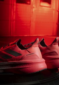 Paire de chaussures de course rouges Adidas Ultraboost avec des rayures noires, placées sur une surface réfléchissante sur un fond rouge.