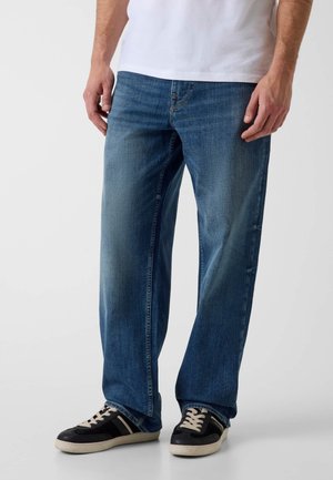 Mann trägt mittelblaue Jeans, schwarz-weiße Turnschuhe und ein weißes Hemd und steht vor einem einfachen hellen Hintergrund.