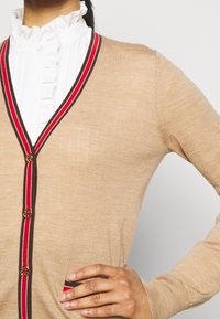 Beige Cardigan mit V-Ausschnitt, roten und braunen Streifenakzenten, goldenen Knöpfen und einer weichen Textur. Mit einer Tasche auf der linken Seite.