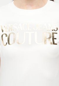 Λευκό πουκάμισο με μεγάλο χρυσό μεταλλικό κείμενο που γράφει "VERSACE JEANS COUTURE" στο στήθος.