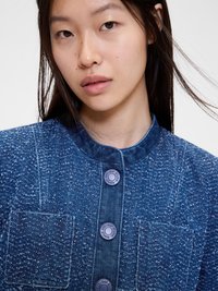 Jachetă din denim în nuanțe de albastru, cu un finisaj texturat, având un guler înalt, închidere cu nasturi în față și un buzunar pe piept.