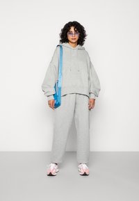 Grauer übergroßer Hoodie und passende Sweatpants, dazu eine blaue Umhängetasche. Pinke und schwarze Sportschuhe sowie eine Sonnenbrille mit blauen Gläsern.