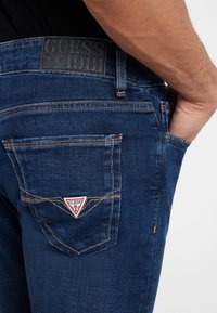 Jeans in denim di colore blu scuro con una finitura testurizzata. Presentano un logo triangolare rosso e bianco sulla tasca posteriore e cuciture arancioni a contrasto.