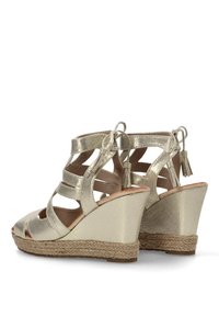 Sandalias de cuña metálicas doradas con diseño de tiras, puntera abierta y acentos de borlas. Suela de cuña texturizada con envoltura de cuerda natural.