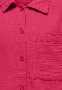 Camisa de tejido ligero y arrugado en un vibrante color rosa, con cuello, seis botones en el frente y un bolsillo de parche.