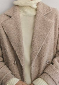 Abrigo de mezcla de lana beige con diseño de doble botonadura, solapas en pico y botones marrones, combinado con un suéter de cuello alto texturizado en color crema.