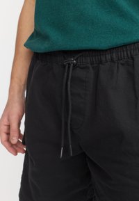 Personne portant un pantalon noir avec une taille ajustable à cordon et une chemise vert foncé, la main reposant le long du corps sur un fond uni.