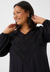 Vestido negro con escote en V y un intrincado detalle de encaje en patrón de chevron. Mangas largas con puños fruncidos. Textura de tela suave.