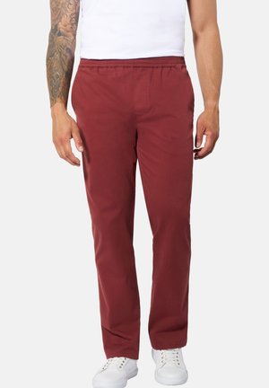 Mann trägt bordeauxrote gerade Hose mit elastischem Bund, weiße Sneakers und ein weißes ärmelloses Oberteil vor einem schlichten hellen Hintergrund.