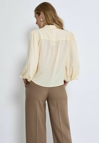 Cremfarbige Bluse mit Puffärmeln und gepunkteter Textur. Mit einem leicht geschwungenen Saum und weichem, leichtem Stoff. Kombiniert mit braunen Hosen.