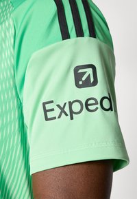 Atletické tričko svetlozelené farby s textúrovaným vzorom, čierne pruhy na ramene a logo s nápisom „Expedia“ na rukáve.