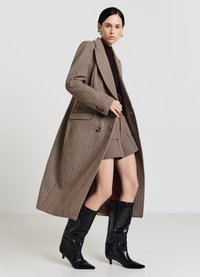 Cappotto doppiopetto in pied de poule marrone con ampi revers, tasche laterali e uno spacco alto, abbinato a stivali neri lucidi alti al ginocchio con punta appuntita.