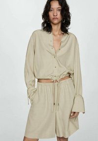 Beige cropped button-up shirt with long sleeves, drawstring bottom, paired with matching shorts. Soft, smooth fabric with a relaxed fit.