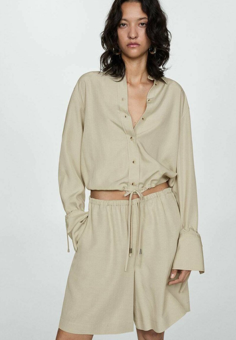 Beige cropped button-up shirt with long sleeves, drawstring bottom, paired with matching shorts. Soft, smooth fabric with a relaxed fit.