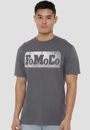 Young man wearing a gray T-shirt with a distressed white "FoMoCo" logo across the chest, standing against a plain background.