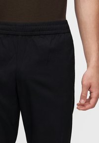 Close-up van een persoon die zwarte elastische taillebroek en een bruin shirt draagt, met de rechterhand ontspannen langs het lichaam.
