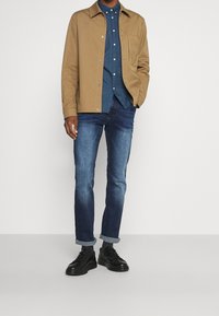 Giacca di cotone marrone, camicia azzurra a pois sotto, jeans blu scuro e sneakers nere. Risvolti dei jeans arrotolati, dettaglio tasca camicia abbottonata.