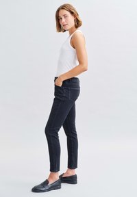 Schwarze Slim-Fit-Jeans mit einem Fünf-Taschen-Design, kombiniert mit einem weißen Tanktop und schwarzen Loafers. Das Outfit zeichnet sich durch eine einfache, saubere Ästhetik aus.