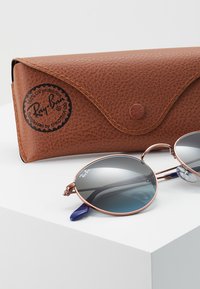 Occhiali da sole Ray-Ban con lenti rotonde sfumate, sottili montature in metallo color oro rosa e estremità del tempio con accenti blu, accompagnati da una custodia in pelle marrone.