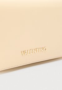 Bež kožna površina s utisnutim zlatnim metalnim logotipom "VALENTINO" blizu donjeg ruba, s finim šavovima uz rub.