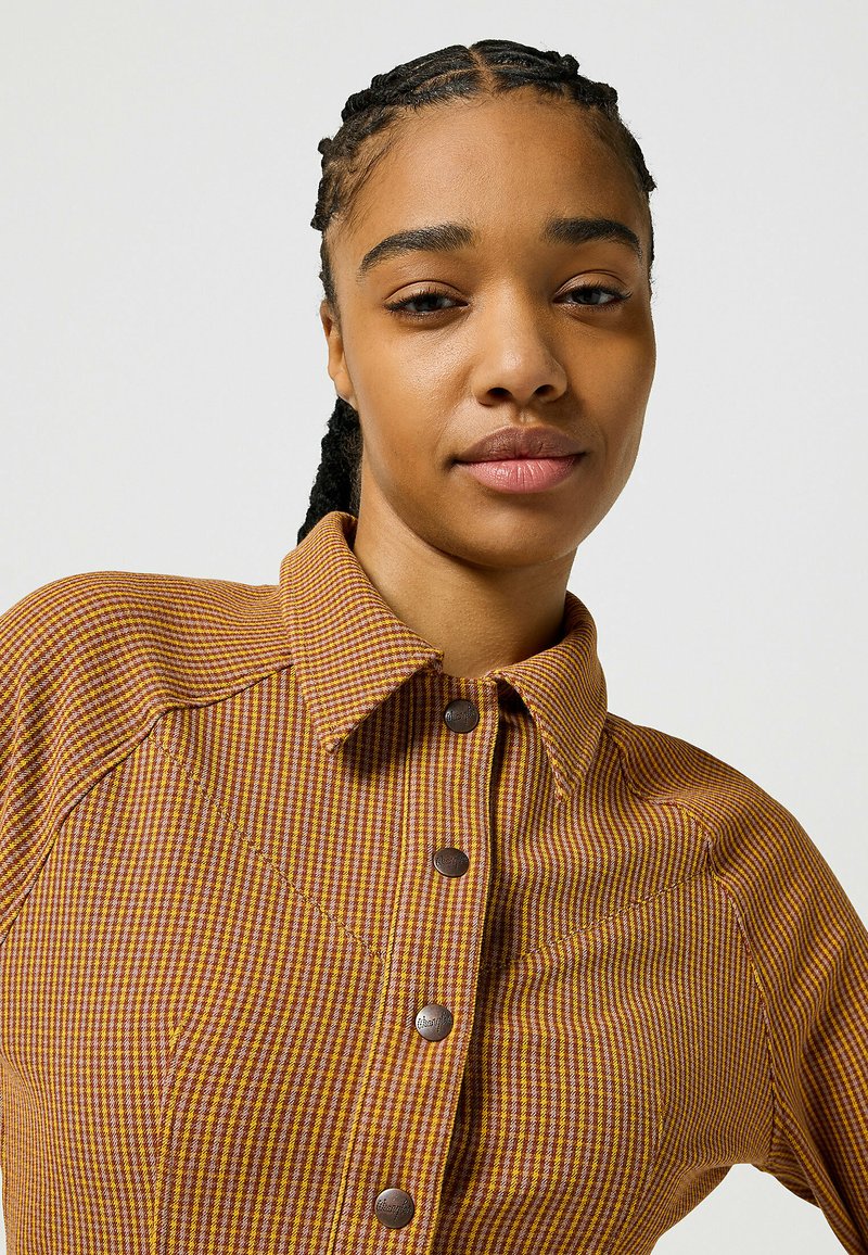 Camicia a scacchi marrone e gialla con colletto strutturato, chiusura con bottoni e tessuto testurizzato. Presenta cuciture a contrasto e cuciture sulle spalle.