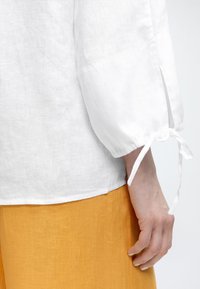 Λευκό πουκάμισο από λινό με μακριά μανίκια και λεπτομέρεια δεμένης μανσέτας, φορεμένο με φούστα από μουσταρδί κίτρινο λινό, σε κοντινή λήψη του μπράτσου και του χεριού.