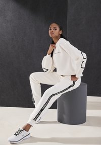 Weißer Sweatshirt und Hose mit schwarzen Logo-Akzenten. Das Design hat Ballonärmel und schmale Beine. Ein Paar schwarz-weiße Sneakers.