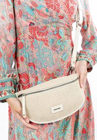 Borsa a tracolla intrecciata beige con una zip liscia, caratterizzata da una forma arrotondata e dettagli in pelle bianca, tenuta contro un vestito a fantasia floreale colorato.
