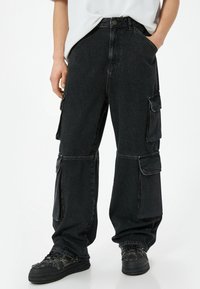 Pantalones cargo de mezclilla negra con un corte relajado, que cuentan con múltiples bolsillos laterales, costuras visibles y una textura ligeramente deslavada.