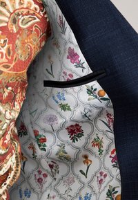 Intérieur de veste de costume marine avec une doublure à motif floral crème, ornée de fleurs colorées et d'une poche à bord noir. Tissu extérieur texturé.